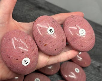 May include: Pink oval-shaped stones with a speckled texture and white numbered stickers. The stones are held in a hand, with several more visible in the background. The stones have a smooth, polished appearance.