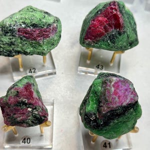 May include: Four rough ruby in zoisite specimens, each mounted on a clear acrylic stand with gold-colored supports. The stones display a mix of green and black zoisite with vibrant red and pink ruby inclusions. Each stand is labeled with a number.