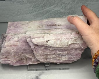 May include: A large, layered crystal formation in shades of pale pink and white. The rough-hewn stone is displayed on a clear stand, showcasing its natural texture and color variations. The crystal is held by a hand.