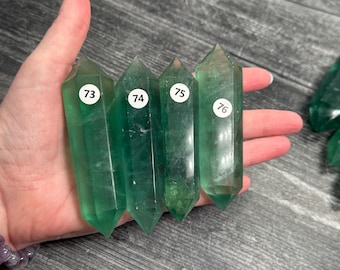 May include: Four green fluorite crystal wands are held in a hand, each with a white sticker displaying a number from 73 to 76. The crystals are translucent with pointed tips. Additional green crystals are visible in the background.