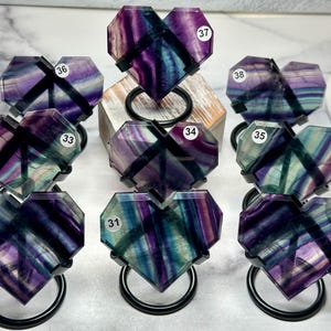 May include: Nine heart-shaped fluorite stones in shades of purple, blue, and green, each with a unique pattern. The stones are displayed on black metal stands. Each stone has a white sticker with a number.