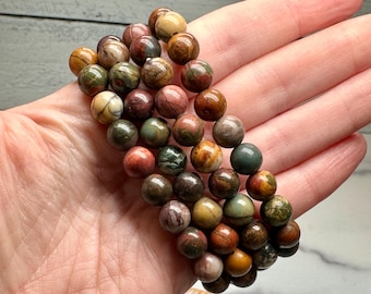 May include: A beaded bracelet featuring round, polished gemstones in shades of brown, green, red, and cream. The bracelet is held in a hand, showcasing the variety of colors and textures of the beads. The beads appear to be approximately 0.3 inches in diameter.