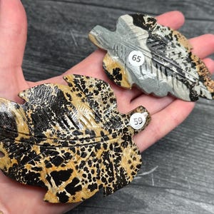 May include: Two carved stone leaf sculptures with intricate details. One leaf is primarily black and tan with a speckled pattern, while the other is gray and tan. Both are held in a hand, showcasing their naturalistic design.