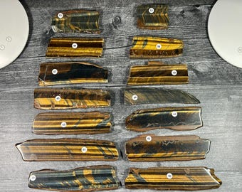 Puede incluir: Una colección de catorce losas rectangulares de piedra ojo de tigre dispuestas en dos columnas. Cada losa presenta distintos patrones de bandas de color marrón dorado y marrón oscuro, con una etiqueta circular blanca numerada del 39 al 52.