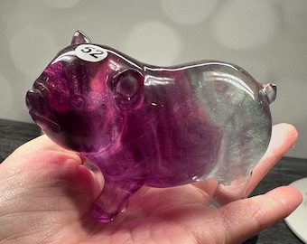 May include: A carved pig figurine made of purple and gray fluorite crystal. The pig has a rounded body, small ears, and a short snout. A white sticker with the number 52 is on the pig's back. The figurine is held in a hand.