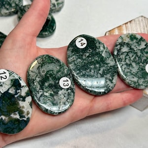 May include: A hand holds several polished oval-shaped moss agate stones. The stones are primarily green and white with a marbled appearance. Each stone has a white sticker with a number on it.