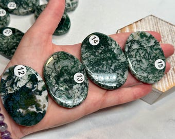 May include: A collection of polished oval moss agate stones featuring deep green and white dendritic inclusions. Each stone is marked with a small white circular sticker displaying a black number, including labels 12, 13, 14, 15, 22, 26, and 27.