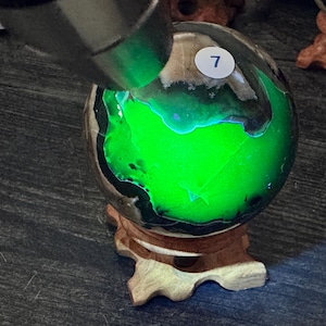 May include: A glowing green sphere with a dark, mottled surface, resting on a wooden stand. The sphere has a bright, internal green light, and a small white sticker with the number 7 is visible on the surface. The sphere is set against a dark wooden background.