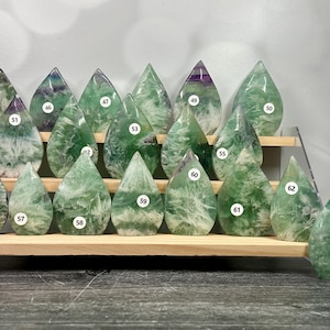 May include: A collection of teardrop-shaped fluorite crystals in shades of green, white, and purple. The crystals are displayed on a wooden shelf, each with a unique pattern and numbered sticker. The crystals vary in size and shape.
