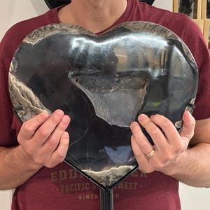 May include: A large, heart-shaped decorative piece made of polished black stone. The stone has a glossy finish with areas of white and gray marbling around the edges. The piece is held in front of a person.