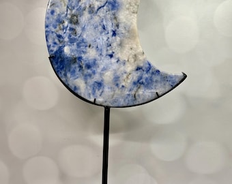 May include: A crescent moon-shaped decorative object with a blue and white marbled pattern, mounted on a black metal stand. The stand has a circular base with cut-out designs. A small white sticker with the number 75 is visible on the moon.