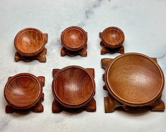 May include: A collection of six wooden display stands in varying sizes. Each stand features a round, bowl-shaped top and a square base with decorative cutouts. The wood has a warm, reddish-brown hue, and the stands are arranged on a light-colored surface.