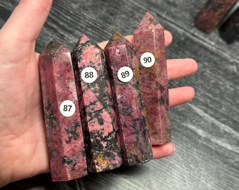 May include: Four pink and black rhodonite crystal points, each with a unique pattern. The crystals are numbered 87, 88, 89, and 90 on white stickers. The crystals are held in a hand.