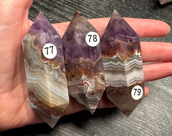 May include: Three amethyst crystal points with layered patterns of purple, brown, and white. Each crystal has a white sticker with a number: 77, 78, and 79. The crystals are held in a hand.