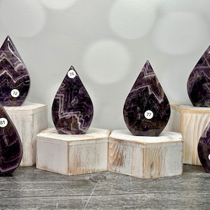 May include: Six teardrop-shaped amethyst crystals with white and purple patterns. Each crystal is displayed on a small, square, white wooden block. The crystals are numbered 72, 76, 77, 79, 81, and 83.