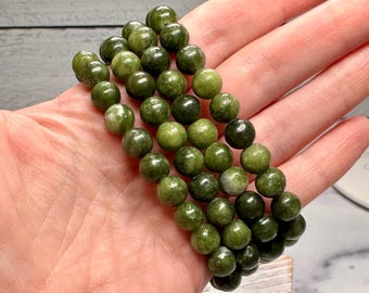 May include: A set of three green jade bead bracelets. The beads are round and have a mottled appearance, with variations in shades of green. The bracelets are displayed on a hand.