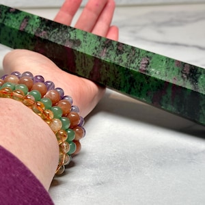 May include: A green and red zoisite crystal obelisk is held in a hand, with a stack of beaded bracelets on the wrist. The bracelets feature a variety of colorful beads, including orange, green, and purple.