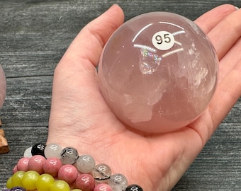 May include: A translucent, pink rose quartz sphere, held in a hand. A white sticker with the number 95 is on the sphere. A beaded bracelet with pink, yellow, and black beads is in the foreground. The background is a dark wood surface.