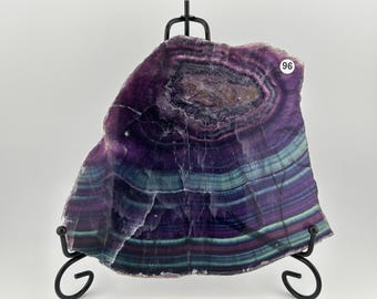 May include: A vibrant, polished fluorite slab, showcasing concentric bands of deep purple, teal, and green hues. The stone is mounted on a black metal display stand, with a small white sticker labeled "96".