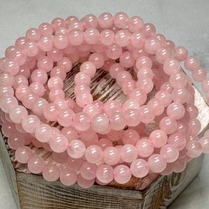 May include: A collection of rose quartz gemstone beads, each approximately 8mm in diameter, strung together to form a bracelet or necklace. The beads are a soft pink color and rest on a wooden box.