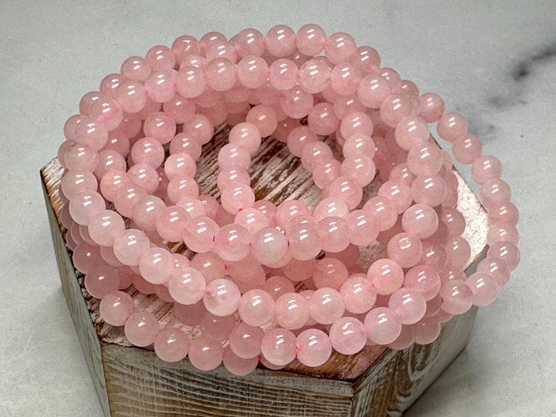 May include: A collection of rose quartz gemstone beads, each approximately 8mm in diameter, strung together to form a bracelet or necklace. The beads are a soft pink color and rest on a wooden box.