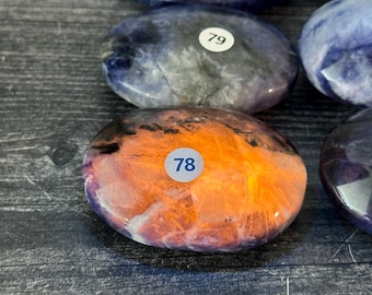 May include: Close-up of several polished stones. One stone is orange and purple with a white sticker labeled "78". Other stones are blue and gray with a white sticker labeled "79". The stones are on a dark wooden surface.