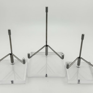 May include: Three clear acrylic display stands with metal supports. Each stand has a square base and a metal rod that extends upwards. The stands are designed to hold items for display.