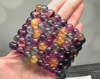 May include: A collection of multi-colored fluorite gemstone beads. The beads are round and vary in shades of purple, yellow, blue, and clear. The beads are strung together, forming a bracelet or necklace.
