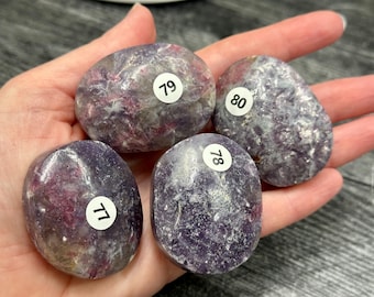 May include: Four polished, oval-shaped lepidolite stones with a mottled purple and grey appearance. Each stone features a small, circular white sticker with black printed numbers 77, 78, 79, and 80, indicating individual inventory identification.