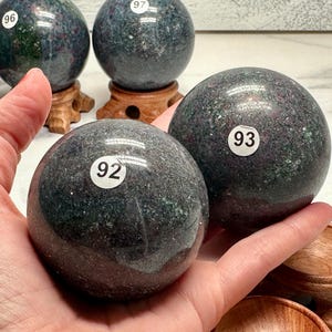 May include: Two dark green and maroon gemstone spheres with white numbered stickers, held in a hand. Other spheres are visible in the background, some on wooden stands. The spheres have a speckled appearance.