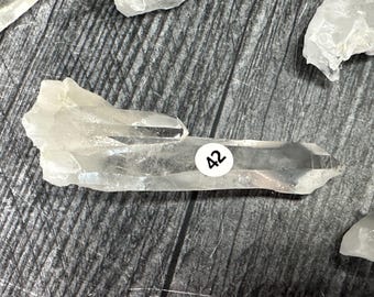 May include: A clear quartz crystal with a pointed tip and a flat, irregular base. A white sticker with the number "42" is affixed to the crystal. The crystal is translucent and appears to be naturally formed.