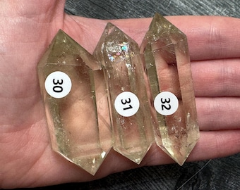 May include: Three double-terminated clear quartz crystals with a pale yellow tint. Each crystal features a white circular sticker with black numbers 30, 31, and 32 respectively. The center crystal displays internal rainbow-colored light refractions.