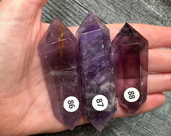 May include: Three double-terminated amethyst crystal points with purple hues and varying levels of clarity. Each crystal features a small white circular sticker with the black printed numbers 86, 87, and 88 respectively, indicating individual pieces.