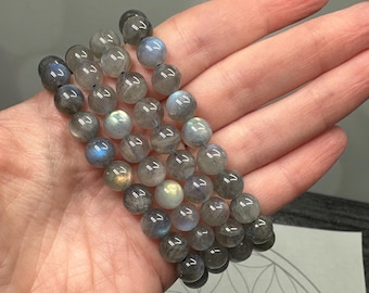 May include: A collection of gray labradorite gemstone bead bracelets. The beads are round and have iridescent flashes of blue, green, and yellow. The bracelets are held in a hand, showcasing the natural variations in color and texture of the stones.