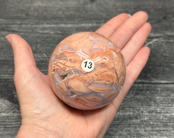 May include: A polished spherical stone featuring a marbled pattern of peach, tan, and light purple hues with visible crystalline fissures. A small white circular sticker with the black number 13 is affixed to the center of the smooth surface.