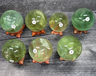 May include: Seven polished green crystal spheres labeled with numbers one through seven. Each translucent orb features internal fractures and natural inclusions, resting securely on a small, carved wooden stand with a dark brown finish.