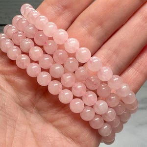 May include: A stack of rose quartz beaded bracelets. The bracelets are made of smooth, round, light pink beads. The bracelets are held in a hand, showcasing the color and texture of the stones.
