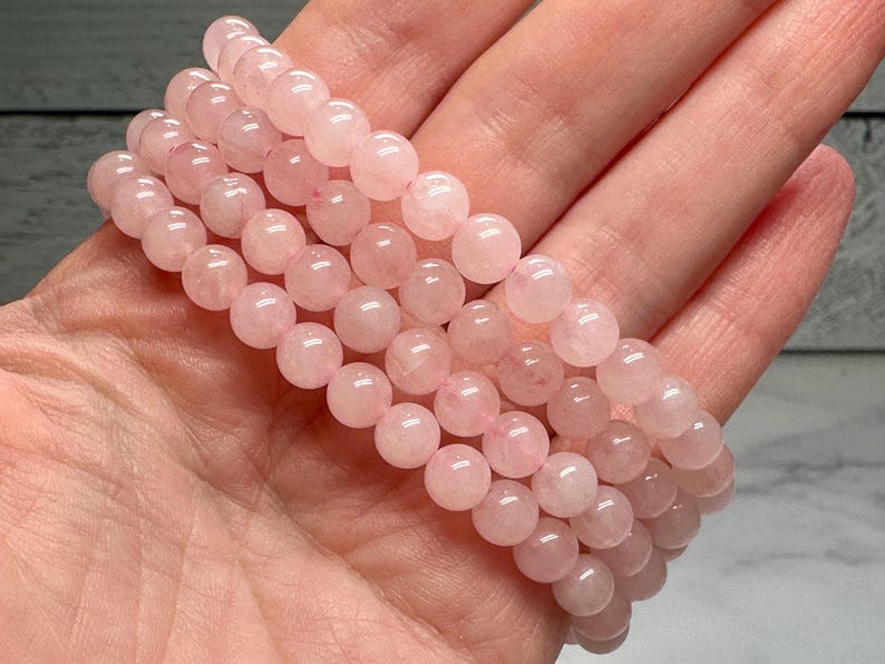 May include: A stack of rose quartz beaded bracelets. The bracelets are made of smooth, round, light pink beads. The bracelets are held in a hand, showcasing the color and texture of the stones.