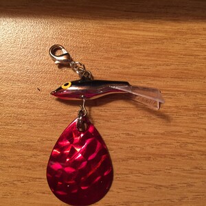 May include: A silver fishing lure with a red and silver blade and a black and yellow fish head. The lure has a clear plastic fin and a silver swivel.