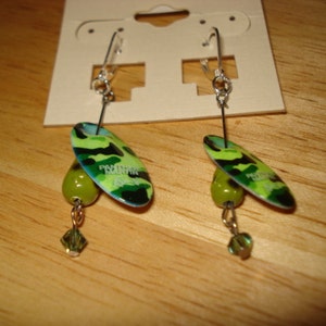 May include: A pair of earrings with green, black, and blue camouflage-patterned pendants. Each pendant has a green bead and a crystal charm hanging from it. The earrings are on a card with the word "Panther" printed on it.