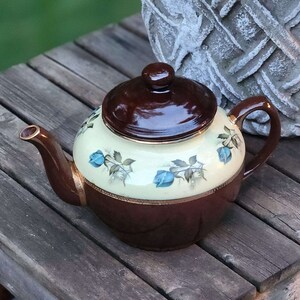 May include: A vintage teapot in brown and cream with a floral pattern, placed on a rustic wooden table. The teapot features a dark brown lid and handle, with gold detailing. A stone planter with red flowers is in the background.
