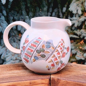 Könnte beinhalten: Eine weiße Keramikkanne mit einem geometrischen und floralen Design in Rot-, Grün-, Blau- und Brauntönen. Die Kanne hat einen runden Körper und einen geschwungenen Griff.