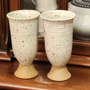 May include: Two white ceramic cups with brown speckles and a textured, ribbed design. The cups have a wide base and a narrow top.