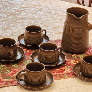 May include: A set of brown ceramic coffee cups and a matching brown ceramic coffee pot. The cups and pot have a textured surface and are arranged on a floral patterned table runner.