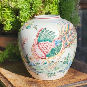 May include: White ceramic vase featuring a hand-painted design of a colourful peacock with green, blue, yellow, and pink feathers. The vase has a round body and a wide opening.