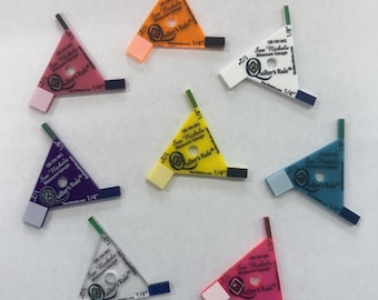 May include: A collection of colorful, triangular quilting gauges. Each gauge features the text "Sue Nickels Measure Gauge" and "Quilter's Rule", with measurements of 1/4 inch and 1/8 inch. The gauges are in various colors, including pink, orange, and blue.