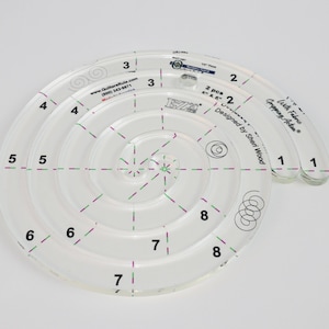 May include: A clear plastic quilting ruler with a spiral design. The ruler has numbers 1 through 8 on the outer edge and is labeled "With Fabric Gripping Action" and "Designed by Sheri Wood".