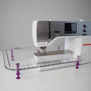 May include: A white sewing machine with a clear plastic extension table. The table has a ruler and four purple feet. The machine has a BERNINA logo on the side.