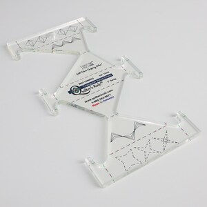 May include: A clear acrylic quilting ruler with a unique triangular swag chain design. The ruler features printed markings and the text "Quilter's Rule" and "Made in America". It is 1/8" thick and has a 3" swag.