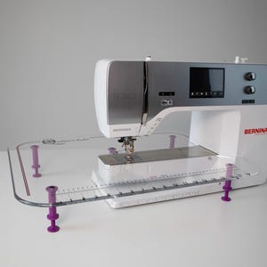 May include: A white and silver sewing machine with a digital display and the brand name "Bernina" in red. A clear quilting ruler with purple supports is in front of the machine. The model number is B 740.